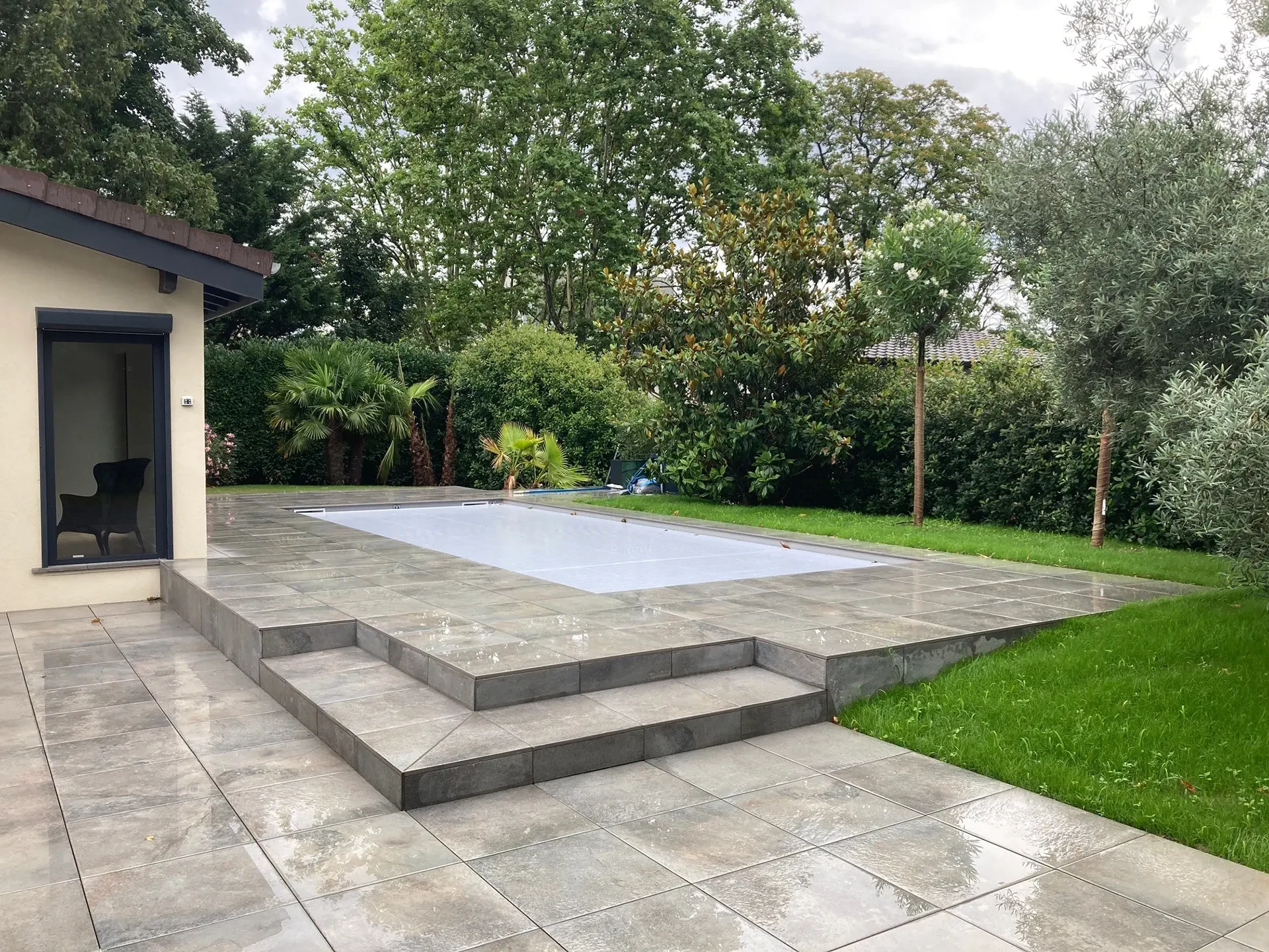 Piscine ovale extérieure avec margelles en pierre naturelle claire, entourée de gazon et végétation méditerranéenne